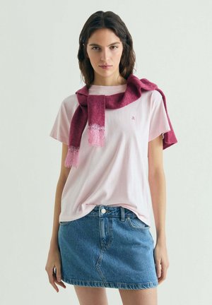 Joven mujer con camiseta rosa claro, falda vaquera y una bufanda rosa oscura sobre los hombros, frente a un fondo liso.