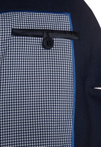 Granatowy materiał z kieszenią w kratkę obramowaną niebieskim kolorem, z pojedynczym czarnym guzikiem. Gładka tekstura z kontrastowymi wzorami.