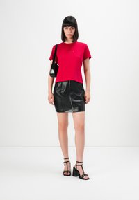 Donna con capelli lunghi e lisci di colore scuro, indossa una maglietta rossa, una mini gonna di pelle nera, sandali con tacco neri e una borsa a spalla nera, mentre è in piedi contro uno sfondo bianco.