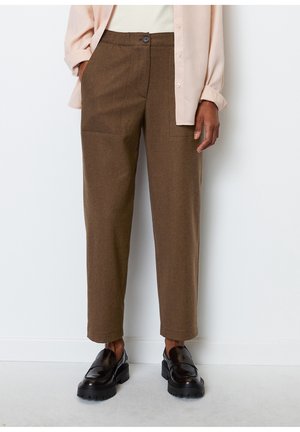 Personne portant un pantalon droit marron avec des poches plaquées, une chemise beige et des mocassins noirs épais, debout contre un mur blanc.