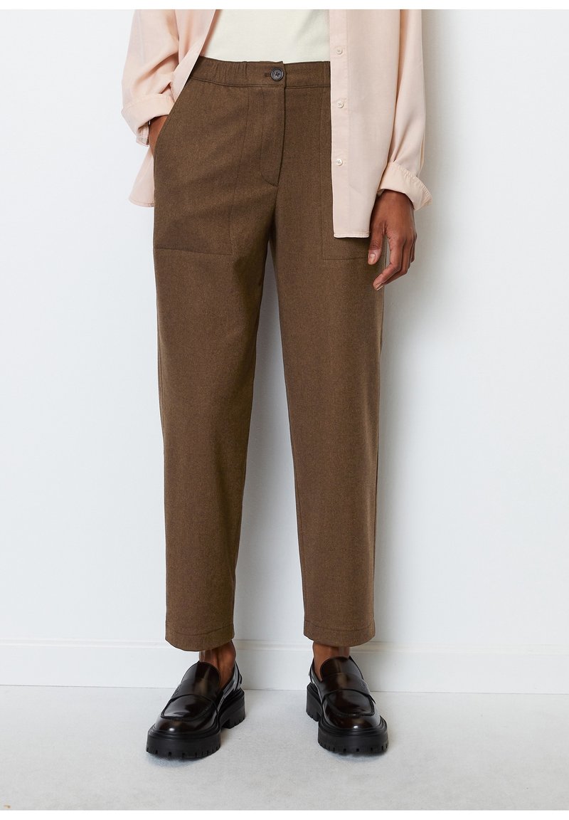 Personne portant un pantalon droit marron avec des poches plaquées, une chemise beige et des mocassins noirs épais, debout contre un mur blanc.