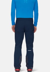Granatowe spodnie outdoorowe o szorstkiej fakturze, z zamykanymi na zamek kieszeniami, wzmocnionymi detalami oraz logiem MAMMUT na dolnej części nogawki.