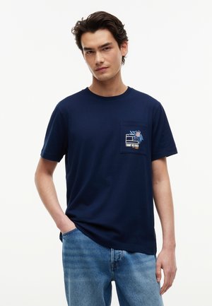 Νέος άνδρας που φοράει μπλε σκούρο T-shirt με μικρό λουλουδένιο και λογότυπο στο τσεπάκι στο στήθος και ανοιχτό μπλε τζιν, στέκεται με το ένα χέρι στην τσέπη.