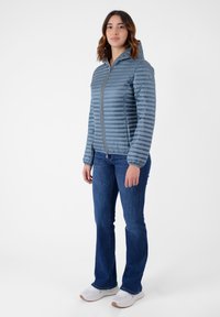 Giacca a vento leggera blu con motivo trapuntato orizzontale, chiusura con zip e cappuccio. Indossata con jeans blu e sneakers bianche.