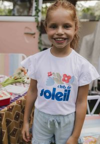 Witte t-shirt met korte gerafelde mouwen, voorzien van een kleurrijk bloemdesign en de tekst "club de soleil" in vetgedrukte blauwe letters. Lichtblauwe shorts.