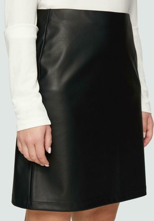 Kobieta w czarnej skórzanej spódnicy do kolan i białej bluzce z długim rękawem, ręce swobodnie opuszczone wzdłuż ciała na jasnym tle.