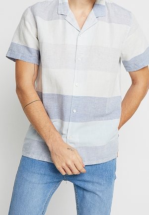 Homme portant une chemise à manches courtes boutonnée avec de larges rayures horizontales bleues et blanches et un jean bleu clair, mains positionnées près de la taille.