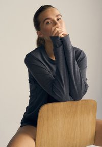 Mørkegrå langærmet skjorte med rund hals og forlængede manchetter, båret af en model, der sidder op ad en lys baggrund.