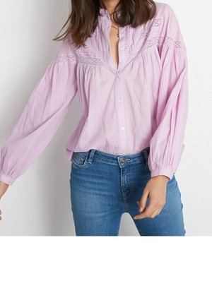 Femme portant un chemisier manches longues violet clair avec des détails en dentelle et un jean bleu, posant avec un bras légèrement étendu.