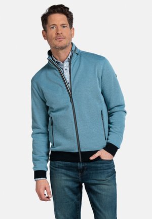 Lichtblauwe zip-up jasje met staande kraag, zwarte accenten en twee met rits gesloten zakken. Gelaagd over een patroonshirt en gecombineerd met donkere jeans.