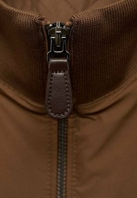 Gros plan sur une fermeture éclair d'une veste en tissu marron avec une languette de tirage en cuir cousue, partiellement fermée au niveau du col.