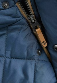 Detail modrého prošívaného saka s zipem a hnědým koženým poutkem na zatahování, kovovým drukem. Hladká textura látky s kontrastním prošíváním.