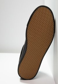 Semelle de chaussure avec un motif de bande de roulement en zigzag marron et des bords noirs, inclinée contre des surfaces blanches dans un coin.