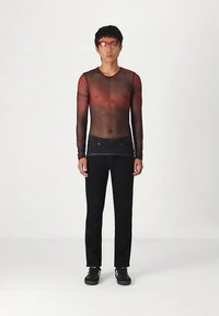 Moški z rdečimi očali, prosojno dolgo-sleeve rdeče in črne majice, črnimi hlačami in črnimi supergami, ki stoji pred enobarvno ozadje.