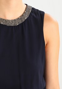 Námořnická modrá bezramenná blůza s perlami na výstřihu. Tkanina se zdá být hladká s mírným leskem, zaměřující se na dekorativní detail límce.