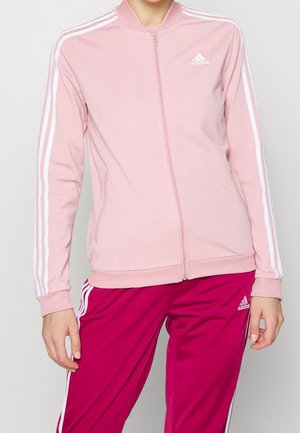 Osoba ubrana w różową kurtkę na zamek marki Adidas z białymi paskami oraz magentowe spodnie dresowe Adidas z białymi paskami.