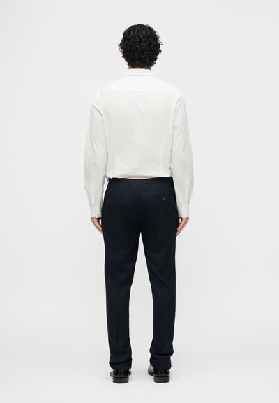 Άνδρας με κατσαρά μαλλιά, φορώντας λευκό πουκάμισο με μακριά μανίκια, μαύρο παντελόνι φόρμας και μαύρα παπούτσια, στέκεται με την πλάτη σε απλό φόντο.