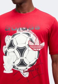 Άνδρας που φοράει κόκκινη μπλούζα Adidas με μεγάλο ποδοσφαιρικό μπάλα, λογότυπο Adidas και έμβλημα Liverpool FC τυπωμένα μπροστά.
