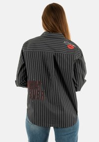 Donna con lunghi capelli castani che indossa una camicia nera a righe con il testo rosso "WHATEVER" e un marchio di rossetto rosso sulla parte alta della schiena, abbinata a jeans blu.