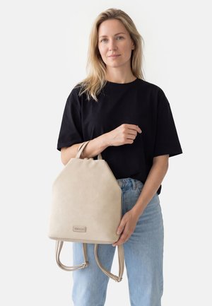 Vrouw met blond haar draagt een zwart shirt en een blauwe spijkerbroek en houdt een beige rugzak met twee bandjes vast tegen een effen witte achtergrond.