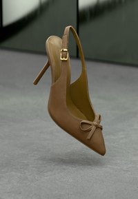 Καφέ δερμάτινη γόβα ψηλοτάκουνα με μυτερή μύτη και μίνι φιόγκο μπροστά, με χρυσό λουράκι, σε ουδέτερο φόντο.