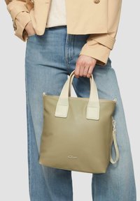 Oliwkowa torba na ramię z kremowymi uchwytami i zamknięciem na zamek. Posiada wytłoczone logo oraz odczepiany pasek. Gładka tekstura.