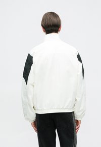 Balta jaka ar melniem akcentiem uz pleciem, piedāvājot augstu apkakli un elastīgas aproces un apakšu. Gluda tekstūra un brīva piegriezuma dizains.