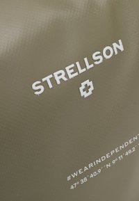 Tessuto verde oliva con superficie testurizzata; presenta il logo in rilievo bianco "STRELLSON" e un hashtag con le coordinate geografiche sottostanti.