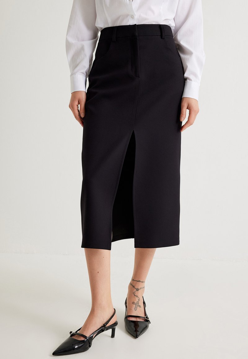 Jupe midi noire avec une fente frontale, design ajusté, texture lisse, associée à des chaussures à talons aiguilles noires et à une chemise blanche à boutons.