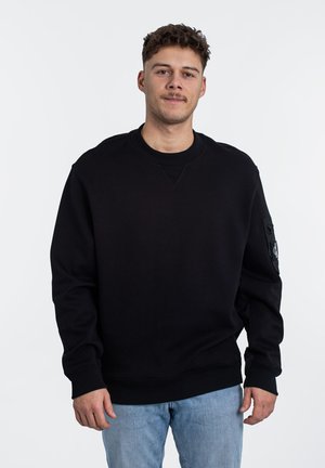 Sweatshirt noir avec un col rond, des poignets et un ourlet côtelés. Présente une poche zippée sur la manche gauche. Porté avec un jean bleu clair.