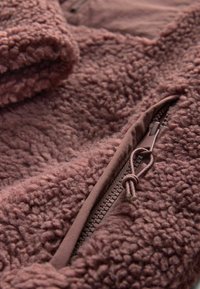 Zbliżenie na jasnofioletową polarową kurtkę, ukazujące fakturowany materiał oraz częściowo rozpiętą kieszeń z sznurkiem do zamka.