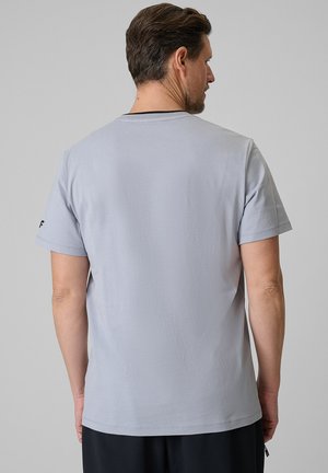 Mężczyzna zwrócony tyłem, ubrany w jasnoszarą koszulkę z krótkim rękawem i czarne spodnie na jednolitym szarym tle.