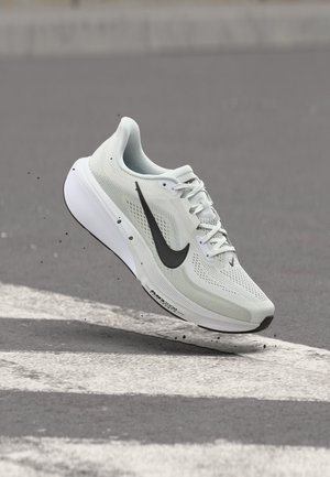 Chaussure de course Nike blanche avec logo swoosh noir, en suspension au-dessus d'un bitume avec des marquages routiers blancs et de petits débris en dessous.