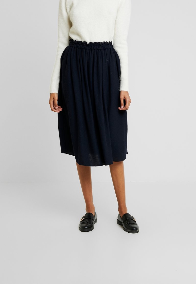 Jupe bleu marine longueur genoux avec une taille élastique froncée, fabriquée en tissu doux et léger. Portée avec des mocassins noirs.