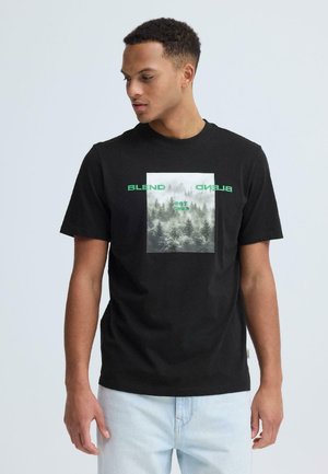 Mand iført sort T-shirt med tåget skovgrafik og grøn tekst "BLEND", der kigger mod venstre mod ensartet baggrund.