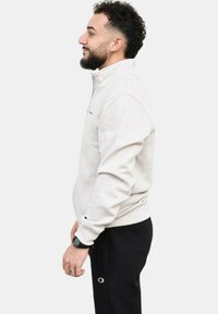 Sudadera cremosa con cremallera, cuello alto, puños acanalados y detalle de logo. Lleva pantalones negros con un logo en el muslo.