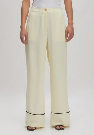 Pantalons large en jaune avec une texture lisse, une fermeture à bouton, et des accents noirs à l'ourlet, associés à un haut ajusté blanc.