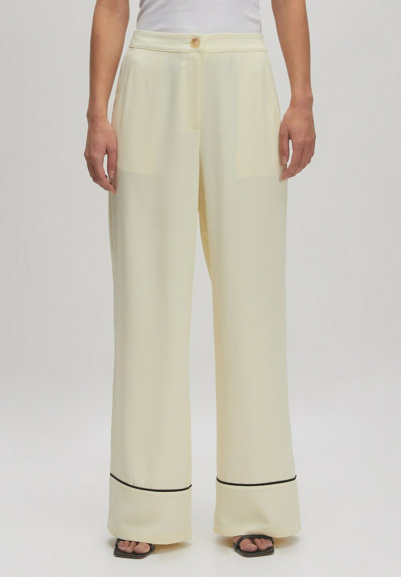 Pantalons large en jaune avec une texture lisse, une fermeture à bouton, et des accents noirs à l'ourlet, associés à un haut ajusté blanc.