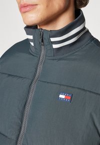 Chaqueta acolchada verde oscura con cuello alto, con cierre de cremallera, cuello a rayas en gris y blanco, y un pequeño parche con logotipo en el pecho.