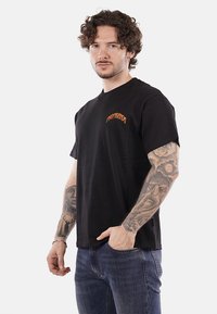 T-shirt nera in cotone con maniche corte, scollatura rotonda e grafica arancione "PROPAGANDA" sul davanti. Indossata con jeans blu.