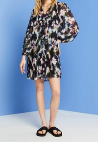 Pleated sheer dress dengan latar belakang hitam dan pola floral beragam warna. Lengan panjang dan desain leher V. Sandal geser hitam.