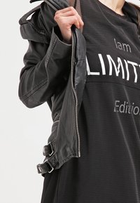 Svart läderjacka med dragkedja fram, strukturerad design, åtsittande manschetter och spännesdetaljer, över en lös svart mesh-topp med tryckt text.