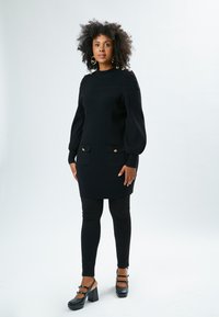 Schwarzes Strickkleid mit langen Ärmeln, gerippter Textur, zwei Fronttaschen, goldene Knopfdetails an den Schultern, kombiniert mit schwarzen Leggings und hohen Schuhen.