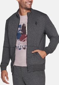 Chaqueta de cremallera de color gris oscuro con textura de punto, puños y dobladillo acanalados, que presenta bolsillos laterales y un pequeño logo en el pecho. Lleva puesta una camisa rosa claro.