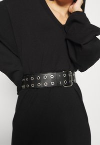 Svart läderbälte med silverfärgade öglor, en rektangulär spänne och en slät yta, bärs över ett svart plagg med vida ärmar.