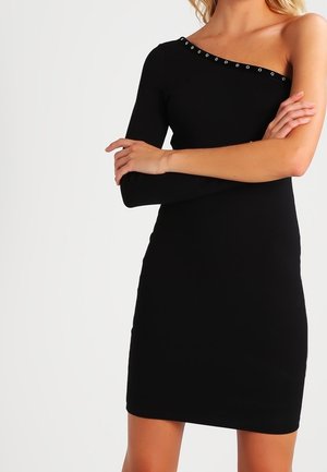 Mujer vistiendo un vestido negro de un solo hombro con una manga larga y detalles de ojales plateados a lo largo del escote.