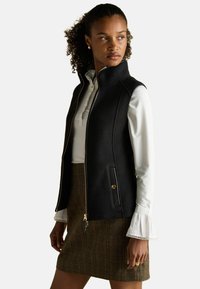 Čierny fleecový vest s vysokým golierom, zipsom a bočnými vreckami. Nosený cez bielu blúzku s volánmi na rukávoch a kockovanou sukňou.