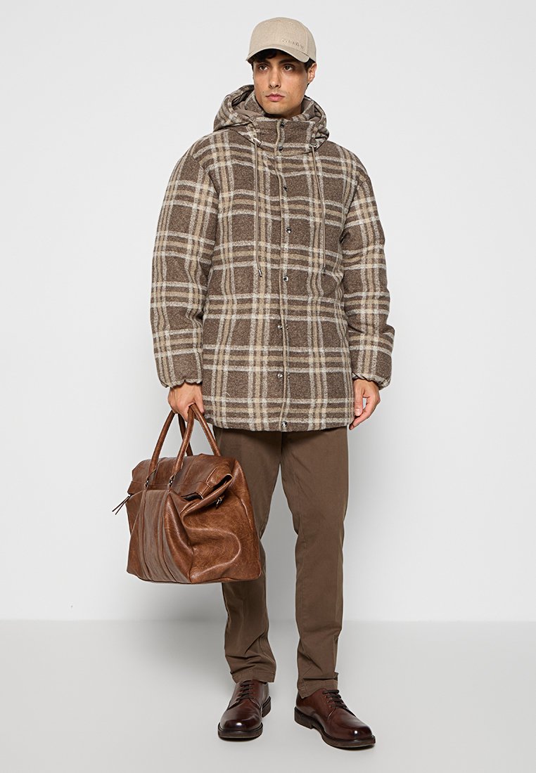 Manteau à carreaux marron et beige avec capuche, fermeture à boutons et cordons; porté avec un pantalon marron et une grande sac en cuir.