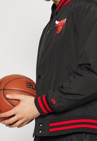 Svart bomberjacka med röda detaljer, med en broderad tjurlogotyp. Ribbstickade muddar och nederkant, mjuk textur. Håller en basketboll.