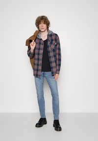 Camisa de franela a cuadros en azul, rojo y verde, con forro de sudadera gris, llevada sobre una camiseta sin mangas negra. Jeans de mezclilla azul claro y zapatos negros.